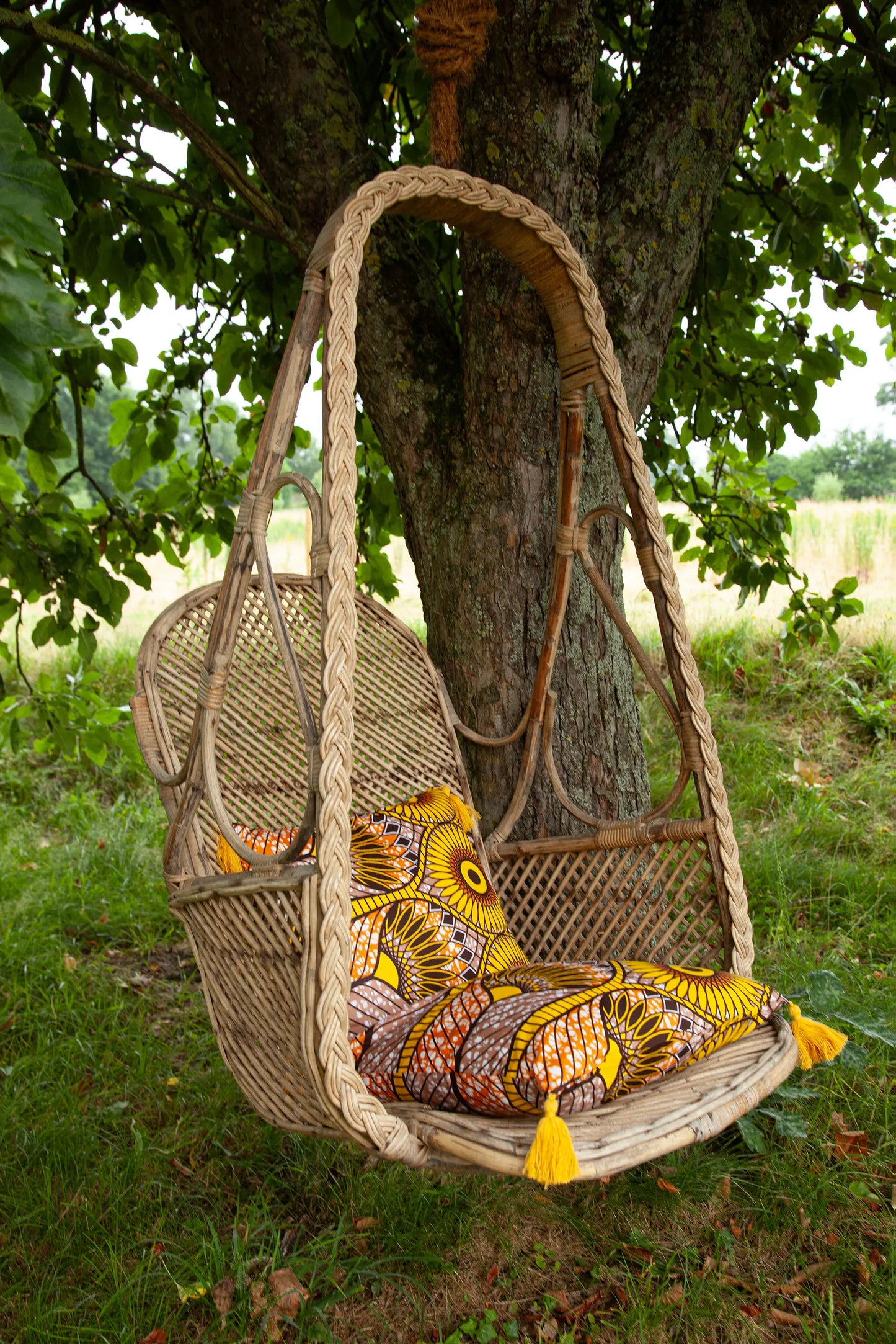 Au jardin, dans une balançoire vintage en osier tressé, deux housses de coussin JUA à pompons en wax jaune et brun. 