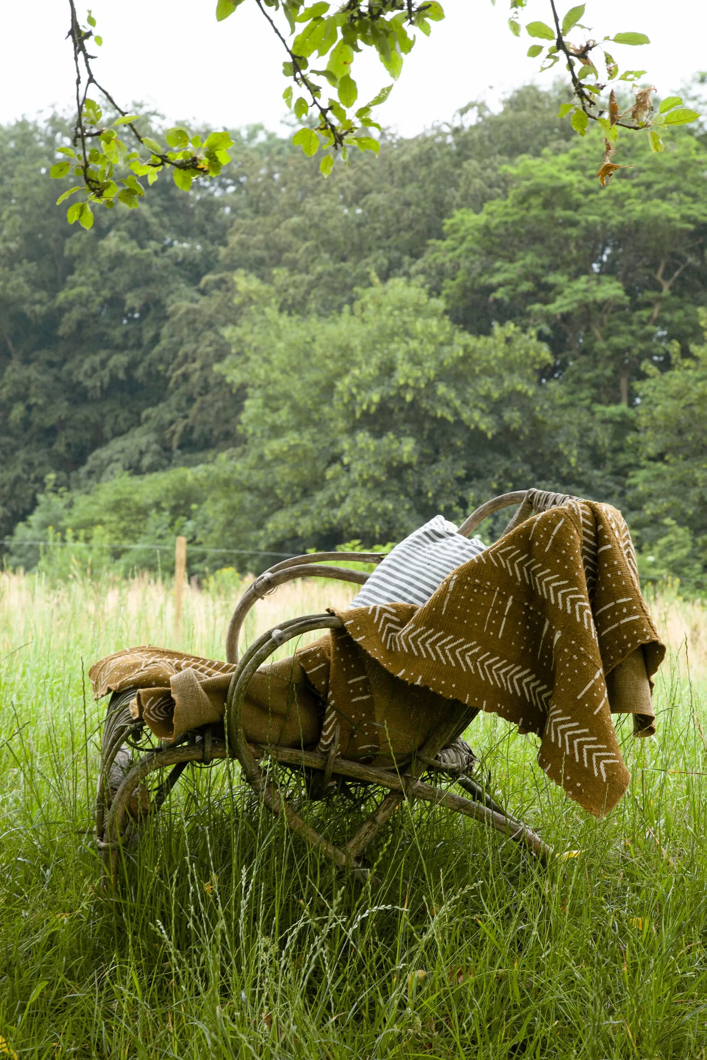 Sur une chaise vintage en rotin, plaid WURA en bogolan ocre de coton bio, fait main et housse de coussin rayée gris et blanc en tissu mossi vintage upcyclé NISHATI. 
Faits main. Studio Matongé