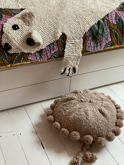 Dans une chambre d'enfant, sur le lit,  une courtepointe FUNTENI par Studio Matonge, un tapis ours polaire en pure laine BEBEAR et au sol, un coussin rond à pompons en laine beige BONBONSI. 
Tous nos articles sont faits main par des artisans qualifiés et passionnés.  
en wax fleuri vert, jaune et ocre et double gaze de coton assortie