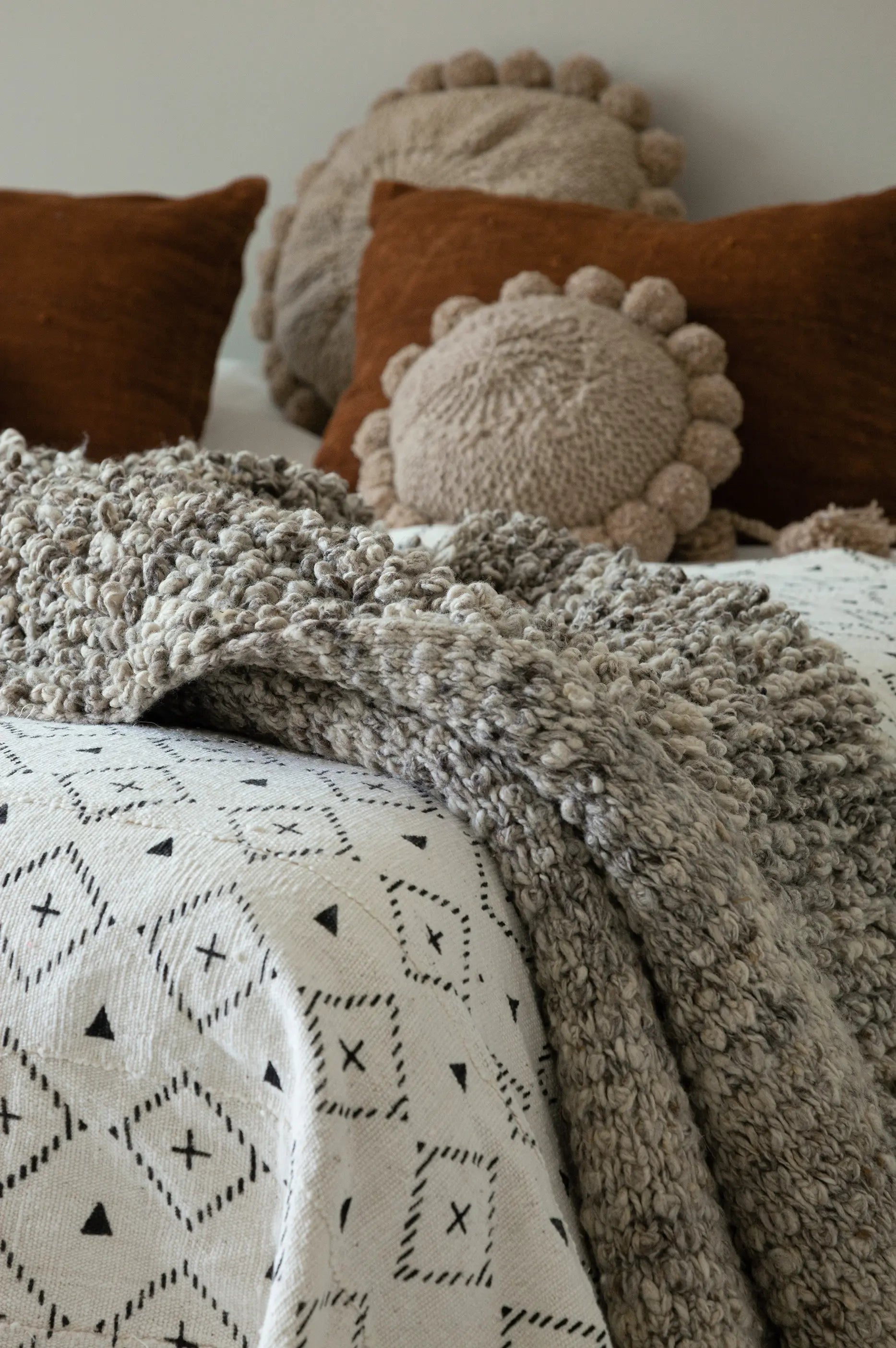 Jeté de lit SUN fait main en bogolan de coton blanc par Studio Matongé, avec le plaid en laine TANGGOOR, les coussins en laine BONBONSI beige et les housses de coussin en bogolan de coton terracotta uni MSINGI. 