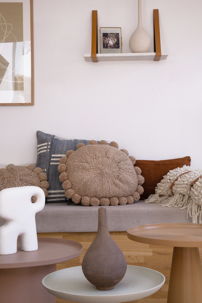 Ambiance arty et détendue pour ce salon avec une grande banquette accueillante couverte de coussins de toutes formes et matières. Les coussins ronds beiges BONBONSI et le plaid en laine écru TANGGOOR en pure laine apportent confort et moelleux. Les housses de coussin en bogolan gris et terracotta NZETE et KIVU réveillent l'ensemble par leurs motifs graphiques ou leur couleur éclatante.  Sur l'étagère murale, le tirage photo Red and Yellow Deep Forest issue de la série Résistance Végétale par Ana Bloom. 