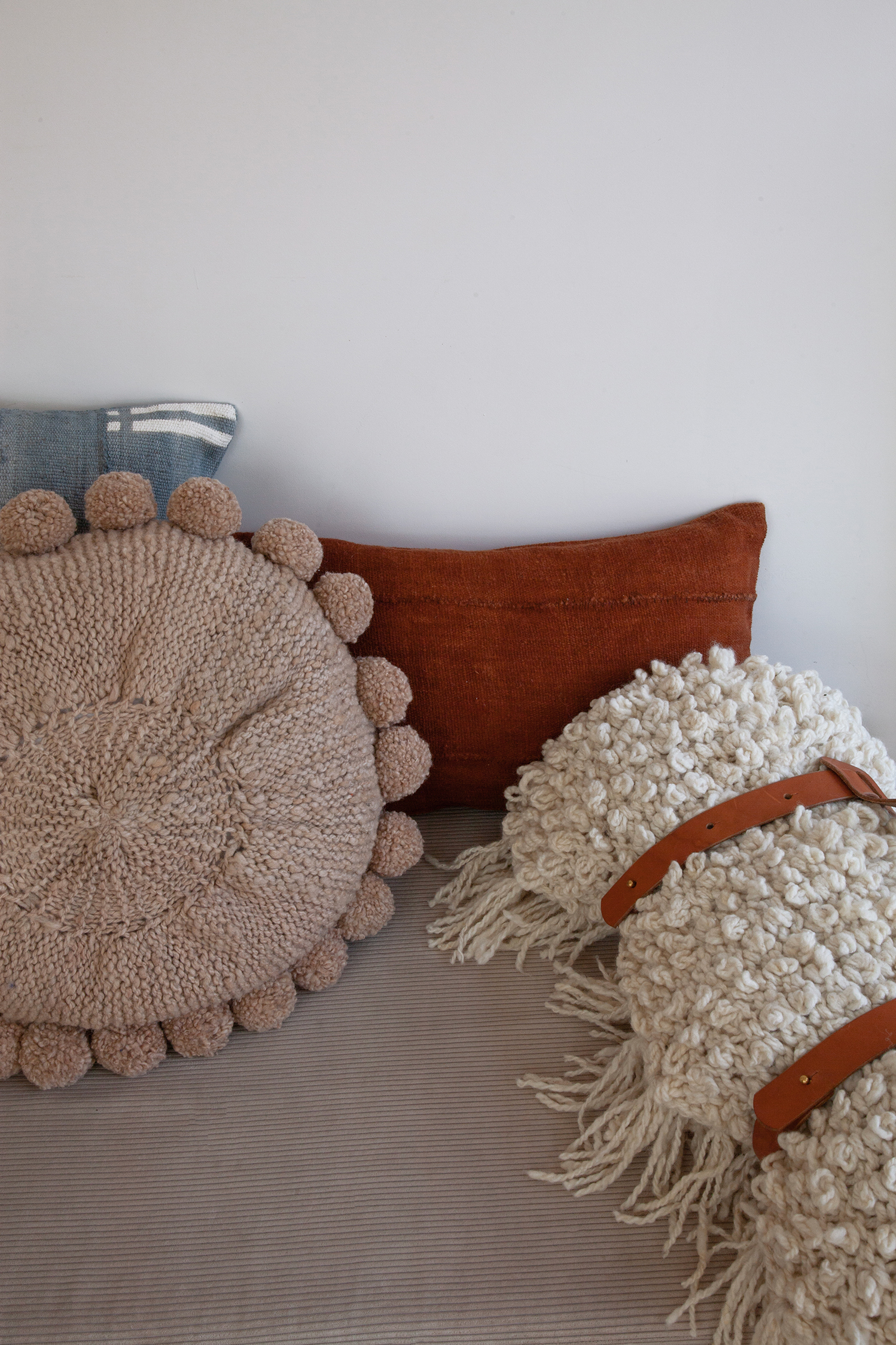 Coussin rond en laine beige à pompons fait main BONBONSI, en combinaison avec les housses de coussins en bogolan gris et terracottta NZETE et KIVU, ainsi qu'avec le plaid en pure laine écrue TANGGOOR et le porte-plaid en cuir TITI par Studio Matongé.