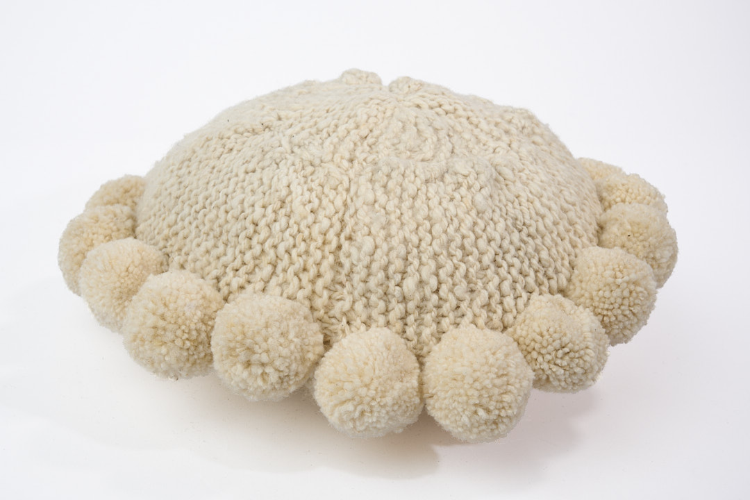 Coussin rond à pompons écru BONBONSI en pure laine mérinos, 30 cm de diamètre. Tricoté main par Kenana Knitters pour Studio Matongé. Disponible en 2 tailles (30 cm et 50cm de diamètre) et 3 couleurs (blanc écru, beige et marron foncé). Insert fourni