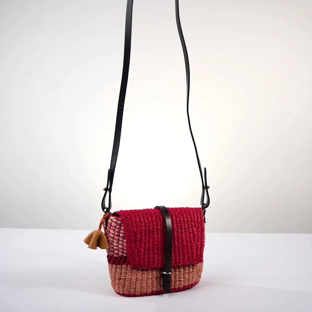 Sac BIKA MINI par AAKS en raphia rouge et saumon. Fabriqué à la main au Ghana, ce modèle compact et pratique est doté d'une lanière en cuir réglable et d'une fermeture à rabat. Porté à l'épaule ou en bandoulière il est parfait pour look bohème et estival.