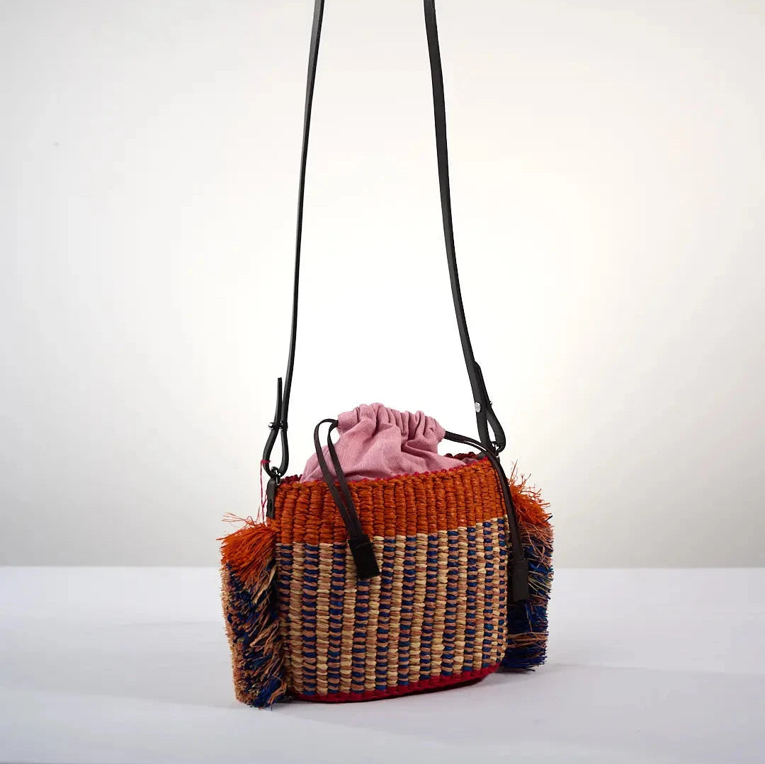 Sac HANA MINI par AAKS en raphia tressé avec un motif original orange et bleu, agrémenté de franges latérales colorées. Fabriqué à la main au Ghana, ce modèle compact et pratique est doté d'une lanière en cuir réglable. Porté à l'épaule ou en bandoulière il est parfait pour ajouter une touche bohème et estivale à toutes vos tenues.