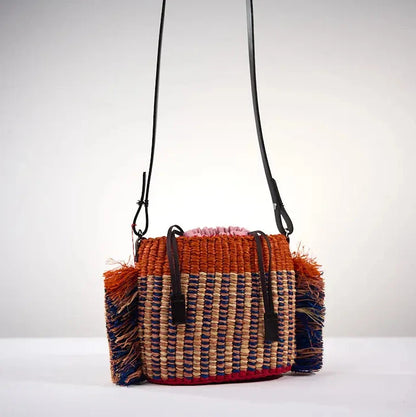 Sac HANA MINI par AAKS en raphia tressé avec un motif original orange et bleu, agrémenté de franges latérales colorées. Fabriqué à la main au Ghana, ce modèle compact et pratique est doté d'une lanière en cuir réglable. Porté à l'épaule ou en bandoulière il est parfait pour ajouter une touche bohème et estivale à toutes vos tenues.