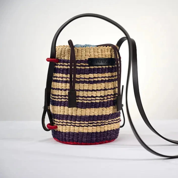 Sac seau LISI par AAKS en raphia avec un motif rayé beige et violet, fabriqué à la main au Ghana. Doté d'une lanière en cuir réglable et d'une fermeture à cordon, il allie élégance et praticité pour un porté à l'épaule ou en bandoulière.