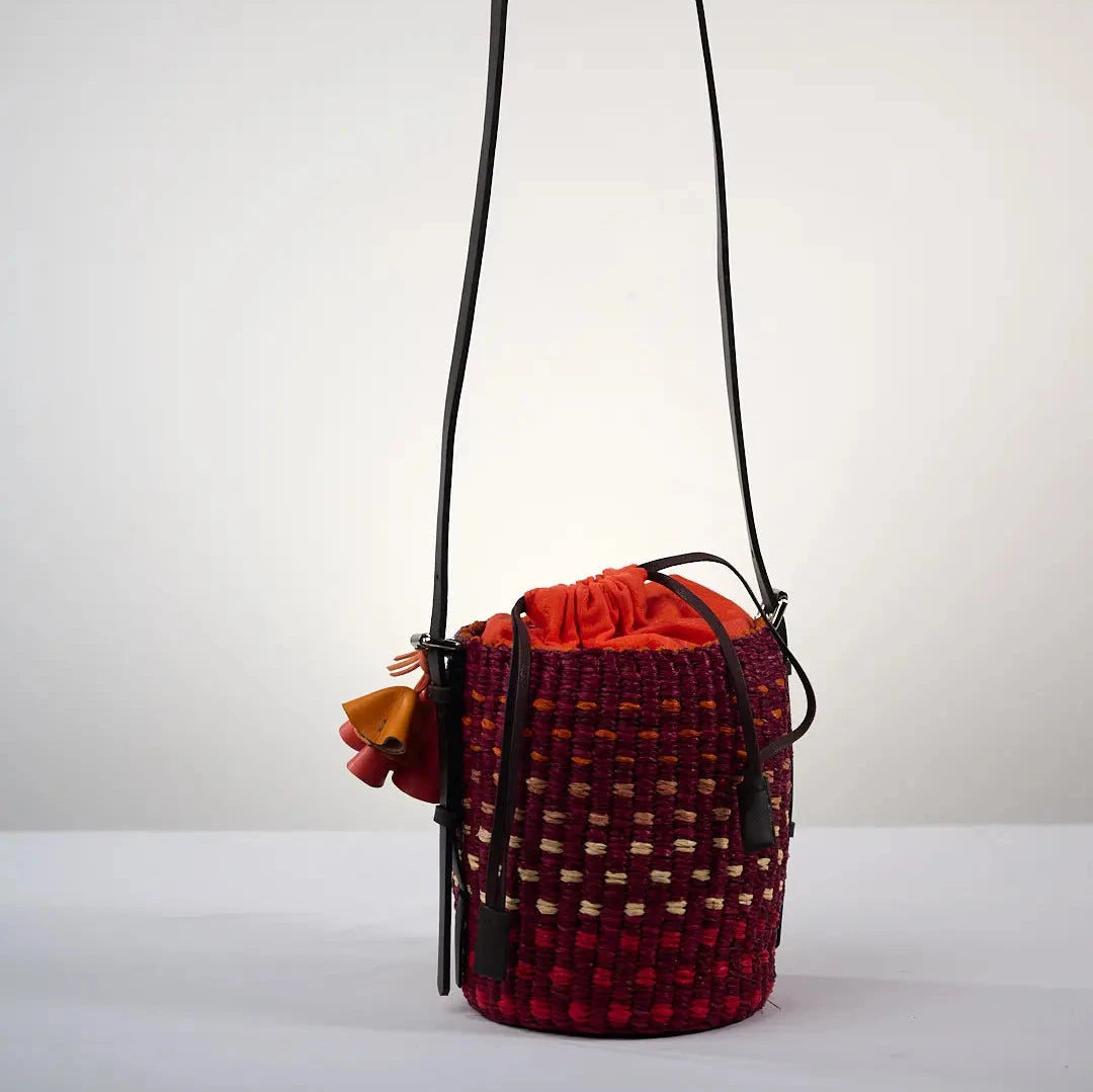 Sac seau LISI par AAKS en raphia avec un motif à pois rouge et orange vif, fabriqué à la main au Ghana. Ce modèle coloré est équipé d'une lanière en cuir réglable et d'une fermeture à cordon, idéal pour un style estival à l'épaule ou en bandoulière.